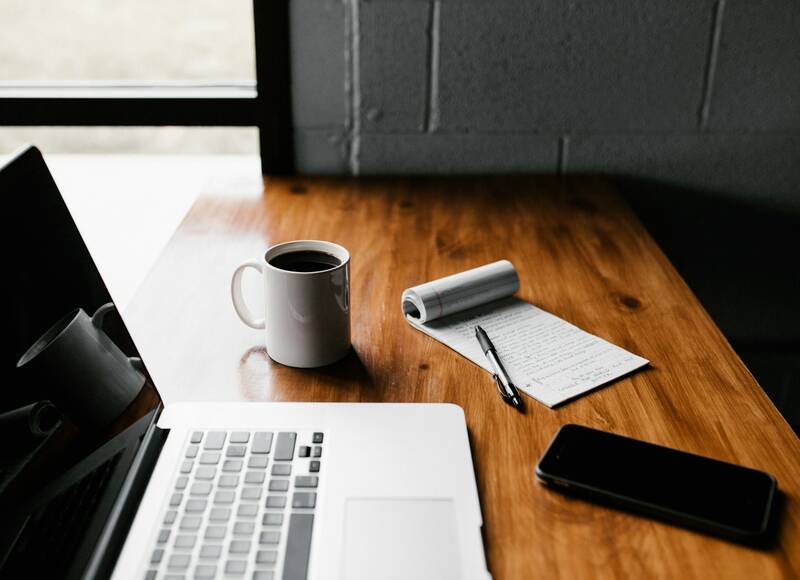 A laptop, a mug of coffee and a notepad on a wooden table